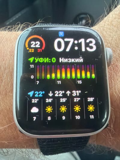 The image displays a close-up of a smartwatch on a person's wrist. The smartwatch screen shows the time as 07:13. It also displays weather information, including a temperature of 22 degrees, with a forecast for 22 and 31 degrees. Below this, there is a graphic with vertical bars and labels "УФИ:0 Низкий" which likely indicates UV index and air quality. Further down, there are hourly weather forecasts with temperatures and weather icons: 22° with clouds at 7, 24° with sun at 8, 25° with sun at 9, 27° with sun at 10, and 28° with sun at 11. The background is blurred, showing the person's arm with hair and what appears to be fabric or a surface. The context of "Wildwood, United States" does not appear to be directly visible or inferable from the image content itself.