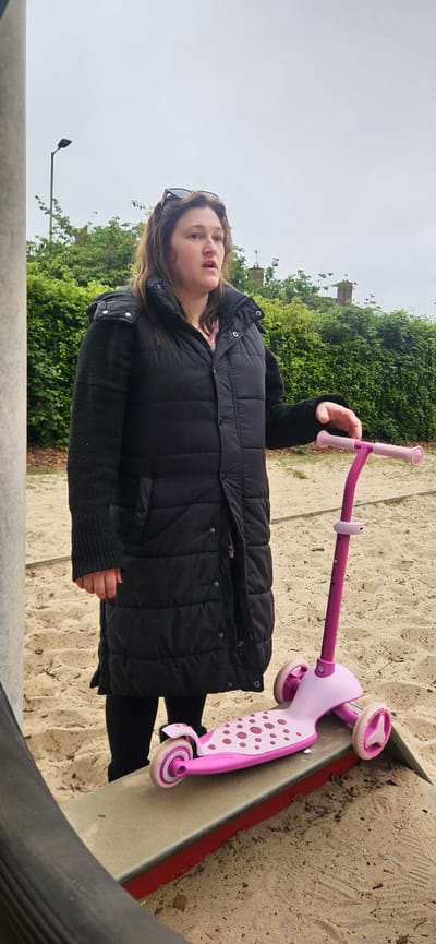 A young woman stands with a pink scooter in a children's playground, which includes a sand pit with a slide feature; the slide is edged in a dark red. The woman is wearing a long black quilted coat with a dark sweater underneath. She has sunglasses perched on her head. The background features lush green bushes and what appears to be a church tower in the distance. A lamppost is also visible in the background. The scene is overcast. The playground location is Norwich, United Kingdom.