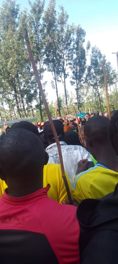 The image shows a group of people gathered in a forested area in Isukha ICHINA, Kenya (latitude: 0.351, longitude: 34.782). The individuals are mostly men, dressed in casual clothing. Some are wearing yellow, red and black shirts. A few people are carrying sticks or walking sticks. The group appears to be in a celebratory mood, with some smiling and laughing. The background shows a dense forest with tall trees and a blue sky. The scene is likely a social gathering or event, with a focus on the group of people at the center of the image.