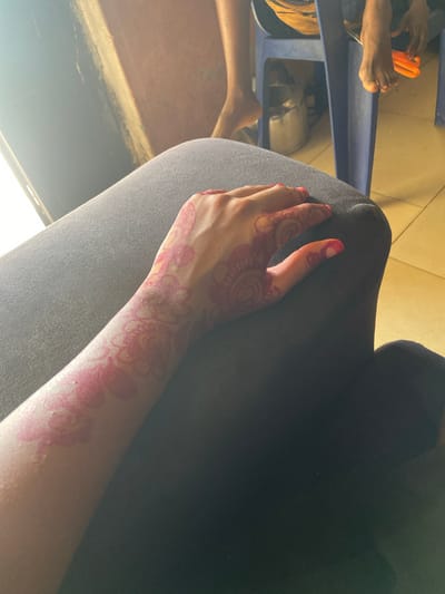 A close-up, high-angle shot shows a woman's left arm resting on a gray fabric surface. The arm features red-pink henna designs, including floral patterns and a large circular design on the back of the hand. Her fingernails are painted red. In the background, out of focus, there are glimpses of other people's legs and feet, and an orange object. The background appears to be a room with tiled flooring. No specific details confirm this is Nagazi, Nigeria.