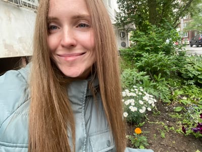 An outdoor, medium shot selfie of a woman with long, light brown hair and blue eyes, smiling. She is wearing a light teal jacket. Behind her, on the right side of the frame, is a garden bed with white daisies, an orange marigold, and some purple flowers. In the background, there are trees and buildings, including what appears to be an apartment complex. The environment suggests a street or residential area in a city. The image shows no explicit indications of Yuzhno-Sakhalinsk, Russia, or any other specific location.
