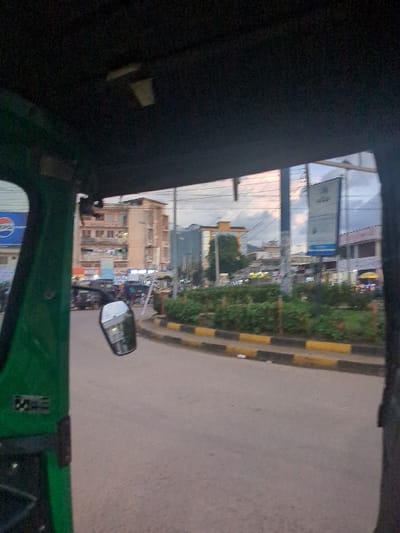 The image shows a view from inside a green vehicle, likely a tuk-tuk, driving along a road in the city of Morogoro, Tanzania. The vehicle's rearview mirror is visible, reflecting part of the street and other vehicles. The street has a black and yellow curb. The road is paved and empty in the foreground. The camera is facing a roundabout, with a planted island in the center. The island is covered in green foliage and has a few trees. Beyond the roundabout, a multi-story building with windows is visible on the left side of the image. On the right, there are several other buildings and signs, with a few people walking on the sidewalk. The sky is partially overcast, with some clouds visible. The scene is characterized by urban traffic, with the tuk-tuk moving along the street. The image captures a typical moment of everyday life in Morogoro, Tanzania.