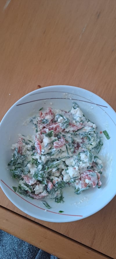 A close-up, overhead shot displays a white bowl filled with a creamy mixture of chopped tomatoes, spinach, and what appears to be feta cheese. The ingredients are finely diced and coated in a white dressing, creating a textured salad. The bowl has a subtle red rim with a thin, dark red line. It rests on a light brown wooden surface, likely a table, with a section of a grey textured fabric visible in the bottom left corner of the frame. The lighting is natural, coming from above and slightly to the left, casting soft shadows. This scene does not contain any people or specific landmarks that would identify its location in Castlebar, Ireland.