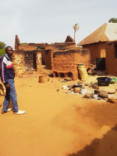 The image shows a scene in Kwal, Nigeria, at the coordinates 9.803 latitude, 8.646 longitude. The scene is an open-air courtyard with a partially destroyed mud brick building on the left side of the image. The building is partially destroyed and has a black, charred exterior, likely caused by a fire. There is a tall, dry palm tree in the background. In the middle of the courtyard is a large, round, mud brick structure, possibly a kiln or an oven, also with a black, charred exterior. The structure has six open vents on its side, suggesting it was used for heating or cooking. There are multiple cooking pots and other utensils scattered around the courtyard, as well as some rocks. A man wearing a blue and red jacket, blue jeans, and white shoes stands in the courtyard, facing the camera. There is a dog in the foreground, partly visible. The scene has an overall dusty, dry, and arid environment. The image shows evidence of past use, particularly the charring and the remains of cooking materials, suggesting that the structure was recently used. The overall tone of the image is one of abandonment or neglect.