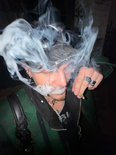 This image, captured in Asnières-sur-Seine, France, displays a close-up of an individual enveloped in a dense cloud of white smoke. The subject, presumably male, is depicted from the upper torso, with his head tilted slightly, exhaling or inhaling from a lit hand-rolled item held between the thumb and index finger of his right hand. His eyes appear closed or heavily lidded.
The individual wears a dark green outer jacket over a partially unzipped black hooded garment, which covers a dark t-shirt with a graphic design. A gray, textured cap is worn backward, with dark sunglasses resting upon it. Visible accessories include multiple silver rings in the left ear, a nose ring, a silver chain necklace, and a black tribal-style tattoo on the throat. Several rings adorn the fingers of the right hand, specifically a large square-faced ring on the ring finger and stacked rings on the middle finger. A black leather strap, potentially from a bag or accessory, featuring metallic studs or spikes, is visible on the left shoulder area.
The background is dark and out of focus, suggesting a nighttime or dimly lit environment. Faint, dark architectural elements, consistent with a decorative metal gate or fence, are discernable in the upper right quadrant.