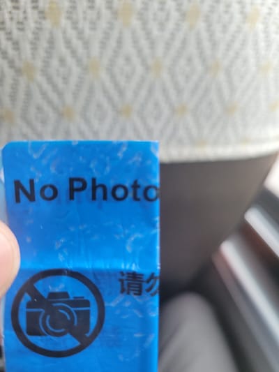 A blue rectangular sign with text and a symbol is partially visible. The text on the upper portion of the sign reads "No Photo". To the lower left, a black circular icon depicts a camera inside a circle with a diagonal line through it, indicating "no photography". To the lower right of the icon, Chinese characters are visible. The sign appears to be made of plastic and has a somewhat textured, crinkled surface. The background is out of focus, showing a light-colored fabric with a diamond pattern and a dark, blurred area. The lighting suggests an indoor environment.