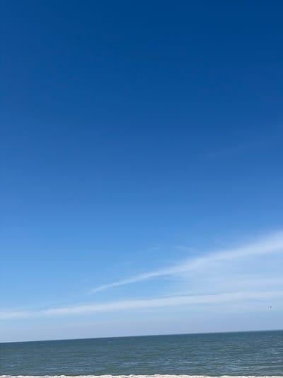 The image captures a serene coastal scene under a clear blue sky. The vast expanse of the sky dominates the upper portion of the frame, transitioning into a thin band of wispy white clouds near the horizon. Below the sky lies the calm, dark blue expanse of the sea, its surface rippled by gentle waves. The distinct line of the horizon separates the water from the sky. In the foreground, a sliver of a sandy beach is visible, appearing light in color. The image is devoid of any people or man-made structures, presenting a natural and undisturbed landscape. The scene is likely a beach in Povarovka, Russia, given the implied context of the location, though there are no explicit visual indicators within the image to confirm this. The overall impression is one of tranquility and natural beauty.