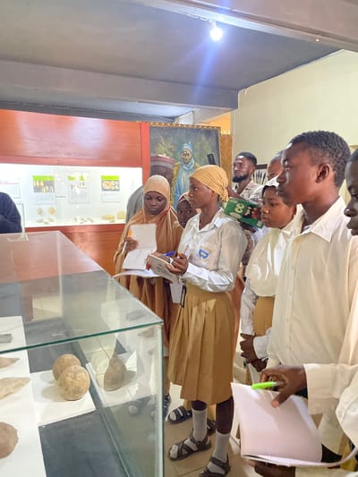 The image shows a group of young people, likely students, observing exhibits in a museum or historical display. The subjects are predominantly of Nigerian descent, dressed in school uniforms. Several individuals are seen taking notes or looking intently at the displays, suggesting an educational visit.
The scene is set indoors, with a museum-like environment. In the background, a large framed painting depicts a figure, possibly historical or religious. Display cases with artifacts are visible, featuring what appear to be geological or archaeological specimens. One display case prominently shows rounded stones or rock samples. Educational posters with text and images are mounted on the walls. The lighting suggests it is daytime, though no specific weather conditions can be inferred.
The activity appears to be a guided tour or a field trip, where students are engaged in learning and documentation. The students' focus and note-taking indicate a serious approach to the educational experience. Their attire, consisting of white shirts and tan skirts or trousers, points to a formal school setting. The overall atmosphere seems calm and studious.
Notable details include the cultural context suggested by the clothing and the painting. The presence of display cases and informative posters confirms the setting as an educational institution or a public display of historical or cultural artifacts. No specific time of day or weather information is discernible. The emotions conveyed are primarily concentration and curiosity. There is some visible text on the educational posters, but it is not legible enough to be transcribed.