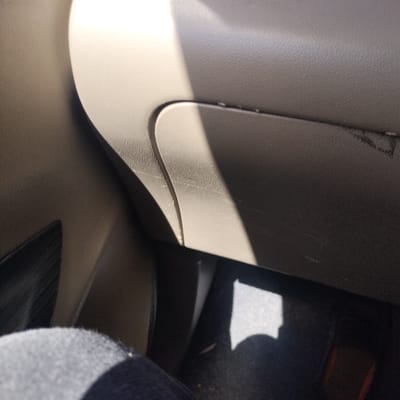 The media file captures the interior of a car, likely from the perspective of a passenger. The central subject is a light beige or tan-colored glove compartment door on the dashboard, featuring a distinct, slightly curved line that suggests its opening mechanism or a design element. Bright sunlight enters the vehicle from the upper right, creating a prominent diagonal highlight across the glove compartment's surface and casting sharp shadows. Faint scratch marks are discernible on the glove compartment.
To the left of the glove compartment, another textured plastic panel, in a darker shade, is visible. The overall color palette of the car interior consists of light beige/tan and darker grey/black tones. In the bottom left corner, a knee or upper leg, covered in dark fabric, is partially visible, indicating a person is seated in the vehicle. The floor area is dark, possibly a car mat or carpet, with a patch of light towards the bottom right.
The scene suggests a sunny daytime setting. No specific activity is taking place beyond being inside a stationary or moving car. There is no visible text in the image. The location context provided is Tanjongkeumala, Indonesia.