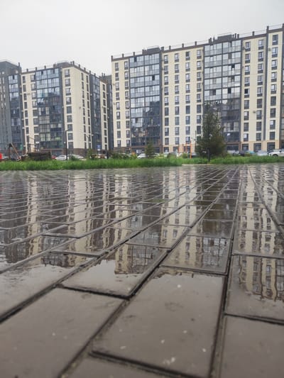 The image depicts a wet cobblestone walkway in front of modern apartment buildings in Prigorodny, Russia. The cobblestones are reflective, mirroring the buildings and the overcast sky. The apartment buildings are tall, with multiple floors featuring numerous windows and balconies. Some air conditioning units are visible mounted on the exterior walls. A strip of green grass separates the walkway from the buildings, and behind the grass, a few cars are parked. The overall impression is one of a residential urban environment after rainfall.