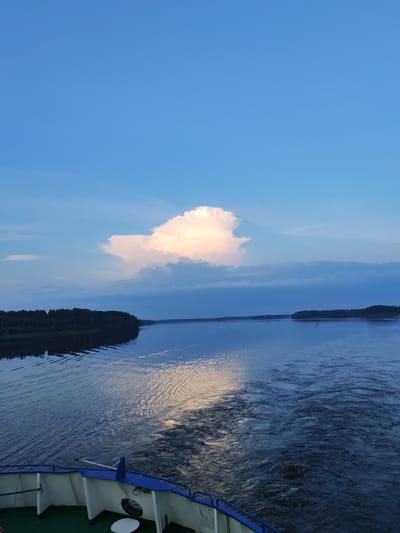 The image depicts a wide river under a twilight sky. The sky is a clear, pale blue with a few wispy clouds, dominated by a large, fluffy cumulus cloud in the upper center, illuminated by the setting sun, casting a warm glow. The river is calm, with gentle ripples reflecting the sky's colors. On the left bank, a dense, dark green forest lines the water's edge. The right bank is further away, also lined with trees, and a small red buoy is visible in the water. In the foreground, the stern of a white and blue boat is visible, with a green deck. The wake of the boat is churning the water behind it. The scene evokes a sense of peaceful travel and the beauty of nature at dusk. The time of day is evening, indicated by the twilight sky and the warm light on the clouds. The location is likely a river in a natural setting, possibly Russia given the contextual information. There is no visible text in the image.