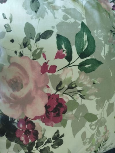 The image is a close-up of a floral fabric pattern. It features several roses in shades of pink and deep red, along with various green leaves and lighter pink or white floral elements. The style of the pattern appears to be watercolor-like, with soft edges and blended colors. The background is a light, pale green or off-white. There are no people, specific objects, or environmental context visible that would allow for the determination of a location in Cēsis, Latvia.