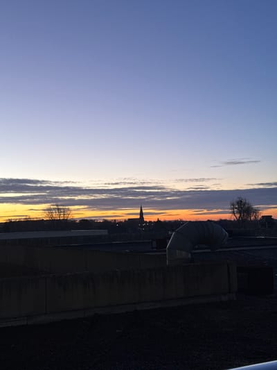 The image presents a panoramic view of an urban landscape in Tourcoing, France, observed from an elevated position, likely a rooftop, during twilight. The sky dominates the upper portion of the frame, displaying a gradient of colors transitioning from a bright orange and yellow along the horizon to light blue and then a deeper purple-blue towards the zenith. Horizontal cloud formations are distributed across the lower sky, reflecting the ambient orange light.
The distant city skyline is silhouetted against the illuminated horizon. A prominent, dark church spire featuring a pointed roof stands centrally within the horizon line, serving as a distinct landmark for Tourcoing. Additional bare trees and various building silhouettes are visible along this distant horizon.
In the midground, on the immediate rooftop from which the image was captured, several dark, rectangular concrete structures or parapet walls are arranged horizontally. To the right of the frame, a large, light grey, segmented flexible ventilation duct curves upwards and then horizontally, consistent with industrial or building HVAC equipment.
The immediate foreground consists of a dark, textured surface, indicative of a flat gravel or asphalt rooftop. No human figures or discernible actions are present in the scene.