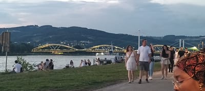 The image depicts a waterfront scene in Linz, Austria, during dusk. In the foreground, a group of people are walking along a path, with a couple holding hands prominently. Behind them, a river is visible, with several individuals seated on the grassy bank. Across the river, a city skyline is illuminated by lights, with two distinct bridges spanning the water. The background features a hilly landscape under a twilight sky. The atmosphere suggests a leisurely evening gathering by the river.