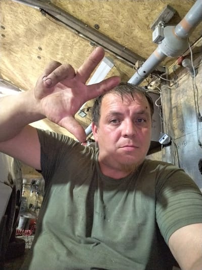 This is a selfie taken by a man indoors, likely in a workshop or garage given the visible tools and industrial-looking ceiling. The man, with short, dark hair and a goatee, is looking directly at the camera with a neutral expression. His right hand is raised, with his index and middle fingers forming a V-shape, pointing towards the ceiling. He is wearing a plain, dark olive green t-shirt.

The background reveals a cluttered environment. The ceiling appears to be made of particle board, with various pipes, wires, and electrical components mounted on it. One large, gray pipe runs diagonally across the upper part of the frame, with orange banding. A bare lightbulb hangs from a wire, providing illumination. In the lower left corner, a portion of a light-colored vehicle or piece of machinery is visible, and further back, what looks like an air compressor hose.

The overall impression is of a functional, possibly dimly lit workspace. There is no indication of the time of day or weather conditions. The man's posture and gesture are ambiguous; they could signify a victory, a greeting, or simply be a casual pose. There is no visible text in the image. The setting suggests a rural or semi-industrial location, and the context provided indicates it is in Novoye Iglaykino, Russia.