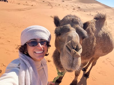 A close-up photograph depicts an adult female and a dromedary camel in a desert landscape near Rissani, Morocco, consistent with the Erg Chebbi dunes. The female, positioned on the left, is smiling directly at the camera. She wears dark-framed sunglasses, a light gray beanie-style head covering, a light beige turtleneck, and a matching light scarf wrapped around her neck. Gold-colored hoop earrings are visible. Her left arm is partially visible, suggesting she is holding the camera. To her right, a brown dromedary camel is positioned, facing partially towards the camera. A light green rope is secured around the camel's head/halter area, visible beneath its chin. The camel's front legs are visible, casting distinct shadows on the sand. The background consists of extensive orange-red sand dunes, marked with numerous footprints and distinct vehicle tire tracks. A clear blue sky is visible in the upper right quadrant. The scene is illuminated by direct sunlight.