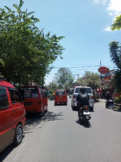 An outdoor, sunlit street scene in Tagtse Tashidingkha, Bhutan, is depicted under a bright blue sky with sparse white clouds. The asphalt road, showing signs of wear, extends from the foreground into the distance, occupied by multiple vehicles in what appears to be a traffic flow or queue.
On the far left, the partial front section of a red minivan is visible, featuring dark tinted windows and silver multi-spoke wheels. Immediately behind it, a bright red minivan is fully in view, displaying white lettering, including "TANGSING," and other stickers on its rear window, with dual exhaust tips visible below. Further down the street, another similar red minivan, marked with a "QUICK 01" sticker, is present. In the mid-ground, a silver SUV is positioned behind the red minivan with the "QUICK 01" sticker.
In the foreground, a person wearing a black helmet and dark clothing with a black backpack is operating a black scooter, facing away from the camera. The scooter has a rear license plate. On the right side of the road, near a curb, another person in a black helmet and dark clothing is stopped on a black scooter.
The right side of the street is lined with buildings, one prominently featuring an oval sign that displays "Lazarro's GRILL & CAFE" in red and orange text. Overhead utility lines traverse the scene. Lush green trees with dense foliage line both sides of the street, casting shadows onto the road surface.