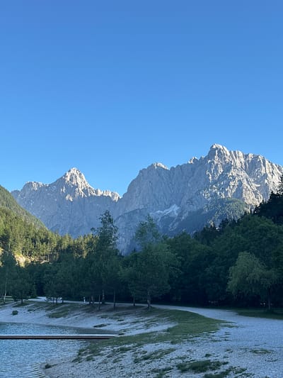 This is a wide-angle, landscape photograph of the Julian Alps, taken from the shore of Jasna Lake in Kranjska Gora, Slovenia. The sky is a clear, bright blue. In the foreground, a body of water with a wooden pier extending into it is visible on the left. The shore is made up of light-colored gravel and sparse green grass. A winding gravel path runs along the shore, bordered by trees. Behind the path and trees, the imposing, rocky peaks of the Julian Alps dominate the background. The mountains are illuminated by sunlight, casting shadows and highlighting their jagged textures. Patches of snow are visible on the higher slopes of the mountains. A dense forest of dark green pine trees covers the lower slopes of the mountains. Two individuals are faintly visible walking on the path in the middle ground. The overall scene depicts a serene, natural environment with a clear, bright day.
