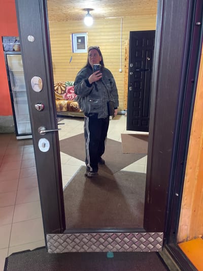 The image shows a person reflected in a mirror, presumably in Картмазово, Russia, although this location information is speculative. The person is holding a phone and taking a photo of themselves in the mirror. The person is wearing dark pants with white stripes and a gray jacket.
The mirror is part of a door. The reflection in the mirror shows a room with wooden walls, a window, and a black door. The floor in front of the door appears to be tiled. The background visible through the door reflection includes part of a sofa with a floral pattern.
