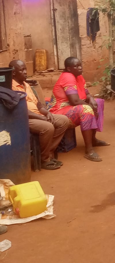 An outdoor scene captures a man and a woman seated on chairs. The man wears a yellow shirt and brown pants with sandals. The woman is dressed in a red top and patterned pants, also sporting sandals. A blue barrel is partially visible, next to a yellow container resting on white material. The backdrop includes a building with hanging laundry. The ground appears to be earthen. Both individuals are looking to the right.