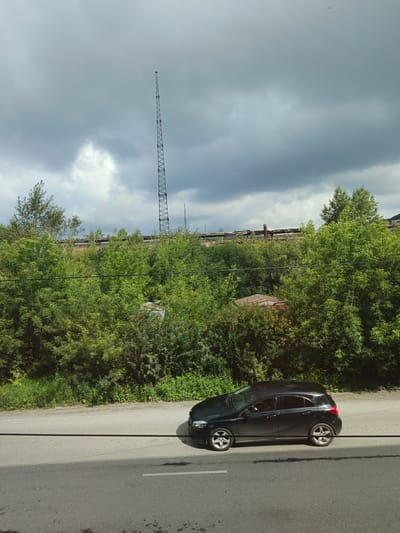 The image depicts a black hatchback car driving on a paved road under a cloudy sky. A tall, lattice telecommunications tower is visible in the background, rising above a dense line of green trees. Partially obscured by the foliage are sections of what appear to be abandoned industrial buildings or structures. A thin, dark cable runs horizontally across the foreground of the image, casting a shadow on the road. The scene suggests a semi-urban or industrial fringe location.