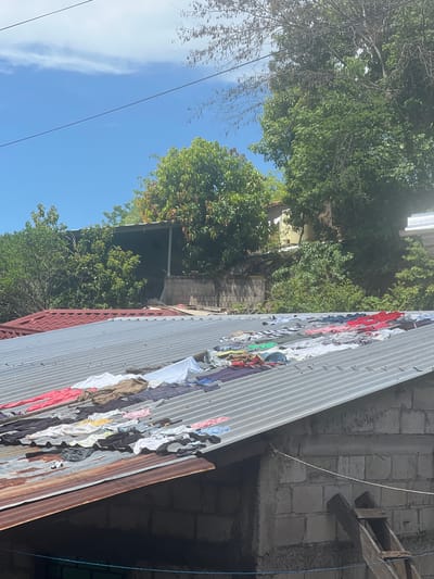 The image depicts an outdoor scene, likely in Amapalita, El Salvador, on a sunny day with a clear blue sky. The foreground is dominated by a corrugated metal roof on which a variety of laundry items are spread out to dry. These items include t-shirts, pants, and other garments in various colors such as red, white, blue, black, and brown. Some of the laundry is neatly arranged, while other pieces are more haphazardly spread.
Behind the primary roof, another roof with red, curved tiles is visible. Beyond the roofs, lush green trees and vegetation cover a hillside. In the background, a building constructed with concrete blocks can be seen, with a wooden ladder leaning against it. Power lines stretch across the top of the image, and faint clouds are present in the sky. The overall impression is one of a residential area, possibly in a rural or semi-rural setting, where clothes are being dried outdoors.