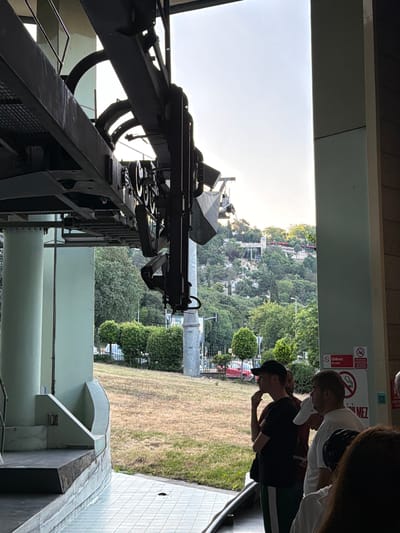 A group of people stand near a cable car station in Eyüpsultan, Turkey, with a lush green hillside in the background. The cable car mechanism is visible overhead. The people are casually dressed and appear to be waiting. The environment is outdoors with dry grass and neatly trimmed bushes. A sign prohibiting entry is visible on a pillar.