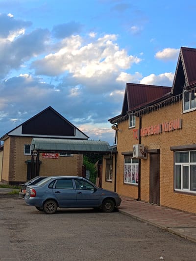 In Zelenchukskaya, Russia, a blue sedan is parked on a gravel lot in front of a two-story brick building. The building has a dark-colored mansard roof with light strings draped across the eaves. A sign with red lettering on the left side of the building reads "АРЕНДА" (RENT) and a phone number. The right side of the building has large orange lettering that reads "КАРАВАН УДАЧИ" (CARAVAN OF LUCK). The sky is blue with scattered white and gray clouds.