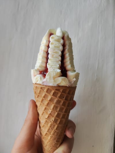 A hand holds a vanilla ice cream cone with red jam swirls against a textured white wall. The ice cream is soft-serve and piped in decorative ridges with visible red jam in between. The cone is a typical waffle cone. The scene is indoors, with the white wall serving as the background. No specific location in Korosten, Ukraine, can be determined from the image.