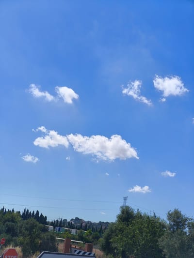 A bright, sunny day in Riba-roja de Túria, Spain, is depicted with a vast blue sky dotted with scattered white clouds. The lower half of the image showcases a landscape characterized by lush green trees and vegetation, with a hint of urban development in the distance, including buildings and power lines. Several small, white, puffy clouds are visible against the clear blue sky. In the foreground, on the left, two red octagonal "STOP" signs are partially visible. The overall scene is tranquil and captures a typical daytime view of the Spanish countryside or outskirts of a town.