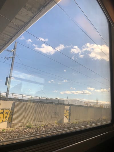 The image provides a view from a train window in Eckwersheim, France, showing a railway environment under a clear blue sky with scattered white cumulus clouds. In the immediate foreground, two parallel railway tracks composed of dark ballast and steel rails are visible. Directly adjacent to the tracks, a long, light-colored concrete retaining wall, characterized by vertical corrugated panels, stretches horizontally across the scene. This wall displays multiple instances of yellow spray-painted graffiti, notably "SFF" or "SSF" tags appearing on both the left and right sections, alongside other faded, darker markings. Sparse, dry vegetation, including weeds and small shrubs, grows along the base of the retaining wall and between the tracks.
To the left, a tall, dark utility pole equipped with electrical insulators and overhead wires is positioned near the tracks. Multiple catenary wires for train power are suspended across the sky, extending diagonally through the frame. In the upper left quadrant, a portion of a concrete overpass or elevated railway structure is visible. Beyond the retaining wall, a metal fence line can be seen, followed by what appear to be low-rise industrial or commercial buildings with light-colored roofs. Distant terrain or structures are faintly discernible on the horizon. The train window itself exhibits minor internal reflections. No individuals are present in the scene.