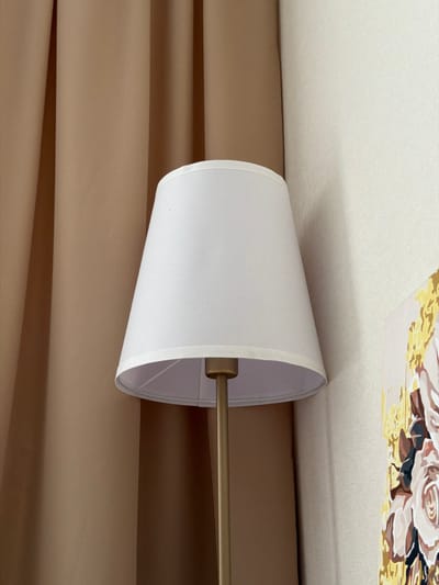 The image shows a close-up of a white lampshade on a golden lamp stand. The lamp is positioned in front of a beige curtain. To the right of the lamp, a section of a painting with floral motifs is visible. The background consists of a textured cream-colored wall. The scene depicts an indoor setting, likely a room, with no visible indications of the specific location within Semiluki, Russia. The lighting is soft, suggesting ambient light or the lamp itself being off.