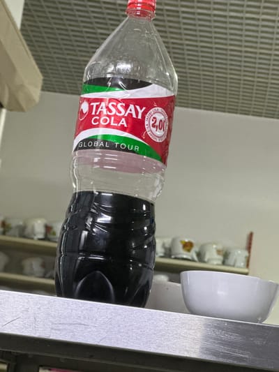 A partially filled bottle of Tassay Cola stands upright on a metal shelf, which is positioned in front of shelves lined with white ceramic cups. The bottle has a red cap and a label that reads "TASSAY COLA" and "GLOBAL TOUR," with a price indicated as "2,00." The liquid inside the bottle is dark brown. The background appears to be an interior space with a tiled ceiling and beige walls. The scene is consistent with a retail or hospitality setting in Kostanay, Kazakhstan.