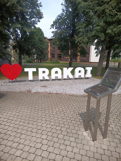 In the city of Trakai, Lithuania, a large red heart is placed next to three-dimensional white letters spelling "TRAKAI." The letters and heart are situated on a bed of white gravel, with a paved walkway in the foreground. To the right of the letters, a metal information stand with a plaque is visible. In the background, trees and buildings can be seen. The lighting suggests it is daytime.
