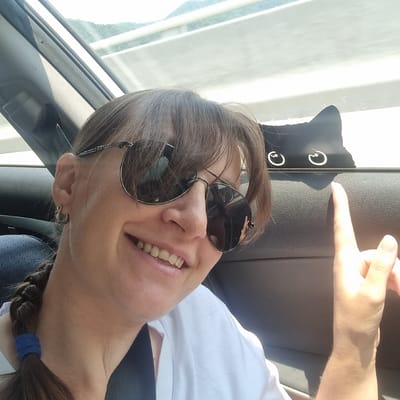 A woman is taking a selfie inside a car. She is wearing sunglasses and a white t-shirt. Her hair is pulled back in a braid. There is a black cat sticker on the car window. The background shows a view of mountains and a road. The image appears to have been taken during the daytime. The location is described as Ucheon-myeon, South Korea.
