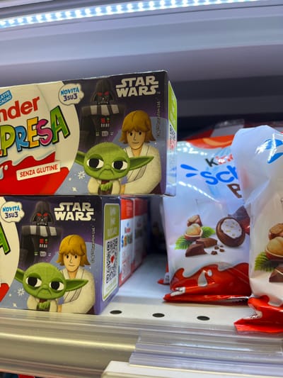 The image displays a retail shelf inside a commercial establishment in Monsummano Terme, Italy. The primary subjects are two stacked Kinder Sorpresa chocolate egg boxes, featuring "STAR WARS" branding. Each box prominently illustrates Darth Vader, Luke Skywalker, and Grogu (Baby Yoda). Visible text on the packaging includes "Kinder Sorpresa," "NOVITÀ 3su3" (New 3-in-3), and "SENZA GLUTINE" (Gluten-free). To the right, multiple white bags containing other Kinder chocolate products, depicting hazelnuts and chocolate pieces, are visible but out of focus. The scene is illuminated by a linear white LED light strip positioned above the shelf. A clear plastic shelf-edge label holder runs horizontally across the bottom of the frame. No individuals are present in the image.