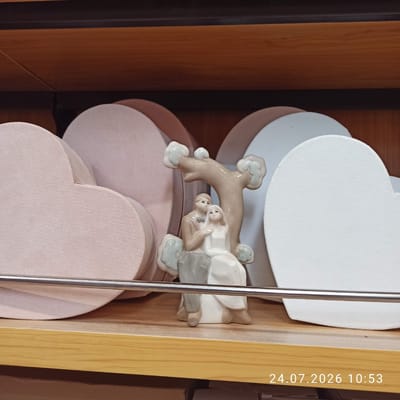 A still life photograph captures a ceramic figurine of a couple sitting under a tree. The figurine is positioned on a wooden shelf, with several large, heart-shaped decorative boxes in pastel pink and white arranged behind it. A metal rod runs horizontally across the lower half of the image, in front of the figurine and boxes. The background appears to be a wooden shelf or display unit. The bottom right corner of the image displays a timestamp: "24.07.2026 10:53". The scene is indoors, likely a retail or display setting. There is no information in the image to suggest a specific location in Modugno, Italy.