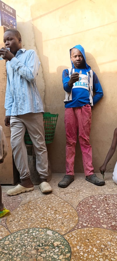 A full-color, vertical photograph depicts two young males and a partial third figure standing against a light-colored wall in Kaura Goje, Nigeria. The setting appears to be an outdoor or semi-outdoor area, illuminated by bright daylight, as indicated by the prominent shadows cast on the wall.
On the left, a young man, likely in his late teens or early twenties, wears a light blue striped long-sleeved shirt and light grey trousers. He is captured in the act of drinking from a small, clear plastic pouch or bottle. His feet are clad in off-white clogs.
To his right stands a younger male, presumably a pre-teen or early teen. He is dressed in a blue and white hooded jacket, which features the word "HILAL" prominently on the front. He wears vibrant pink or red pants and dark, textured slip-on shoes. He holds a small, light-colored object in his right hand, looking down towards it.
A partial view of a third, smaller figure, a child, is visible on the far right, wearing a white garment and holding a small green object. Behind the two main figures, a green plastic basket is visible.
On the upper left side of the image, a sign is affixed to the wall. Visible text on the sign includes "C MAN" at the top, followed by "24" AC DC CEILING FAN," and partially visible text indicating "SWEEP 800MM."
The ground features a distinctive terrazzo floor patterned with large, irregular shapes in shades of light brown, green, and reddish-brown, separated by dark lines. The scene conveys a casual, everyday moment in a local setting.