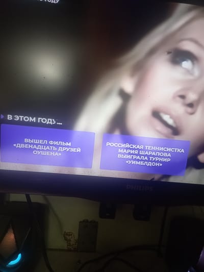 This media file is a photograph of a television screen displaying content related to sports and film. The screen shows two purple text boxes with Russian text. The upper box reads, "РОССИЙСКАЯ ТЕННИСИСТКА МАРИЯ ШАРАПОВА ВЫИГРАЛА ТУРНИР «УИМБЛДОН»" which translates to "Russian tennis player Maria Sharapova won the Wimbledon tournament." This suggests a news or sports highlight.

The lower box reads, "В ЭТОМ ГОДУ... ВЫШЕЛ ФИЛЬМ «ДВЕНАДЦАТЬ ДРУЗЕЙ ОУШЕНА»" which translates to "This year... the film 'Ocean's Twelve' was released." This indicates a segment about film releases. The background of the screen appears to be a blurred image of a woman's face, possibly associated with the content displayed.

The lower right corner of the image shows part of a Philips television. The overall scene is dimly lit, and the screen itself is the primary source of light. There are some dust particles visible on the screen. The image is oriented vertically. The context provided places the scene in Maloye Lugovoye, Russia.