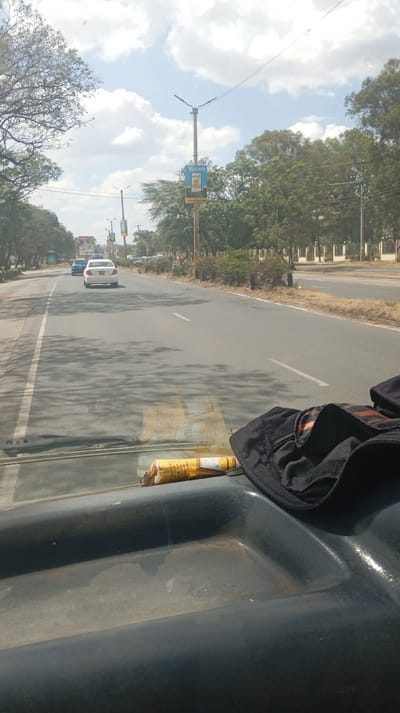 A view from inside a vehicle shows a long, straight road lined with trees and occasional streetlights. A white car is visible ahead, driving in the same direction. The road surface is mostly gray, with some areas darker, possibly due to shadows. A broken cigarette pack and dark garment sit on a bluish-gray dashboard. Buildings and a wall-like structure are seen in the distance, bordering the road on the right. The sky is bright with scattered clouds.