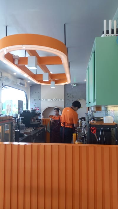 A horizontally oriented medium shot captures the interior of a coffee shop, presumably a "Punthai Coffee" location in Plai Phraya, Thailand. An individual, likely a barista, wearing an orange shirt and apron is standing behind a counter, possibly preparing a beverage. A reflection of the barista and their workspace is visible in a reflective surface. The coffee shop appears to be modern, with orange and white decor, including a unique orange rectangular frame with white cube accents hanging from the ceiling. The room is illuminated by overhead lighting. Various coffee-making equipment and supplies are visible on the counter. A ventilation unit is mounted on a wall. The photo is taken at eye-level.