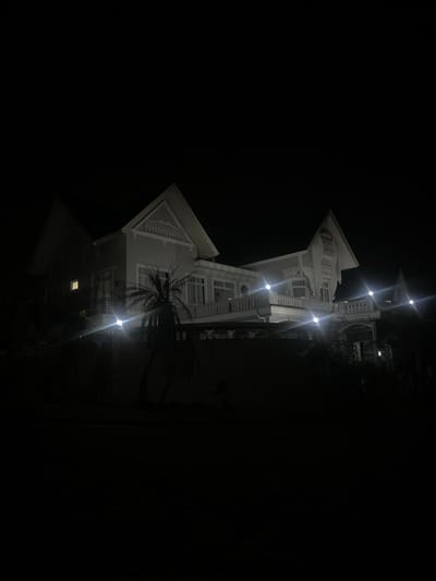 The image displays a multi-story residential structure, identified as located in Batulawang, Indonesia, at night. The house is predominantly light-colored, featuring a complex architectural design with multiple gabled roofs, ornate eaves, and several balconies or terraces adorned with white balustrades. Prominent white outdoor floodlights are affixed to the facade and perimeter of the building, generating significant lens flare. One interior window on the left side of the structure emits a warm, yellow light. A tall palm tree stands in the immediate foreground on the left, partially obscuring the lower section of the house. Dark foliage and a low wall are visible at the base of the property, separating it from the completely dark foreground, which suggests ground level or a roadway. The sky above is uniformly dark, indicating nighttime conditions. No individuals are visible in the scene, and no specific actions or interactions are depicted.