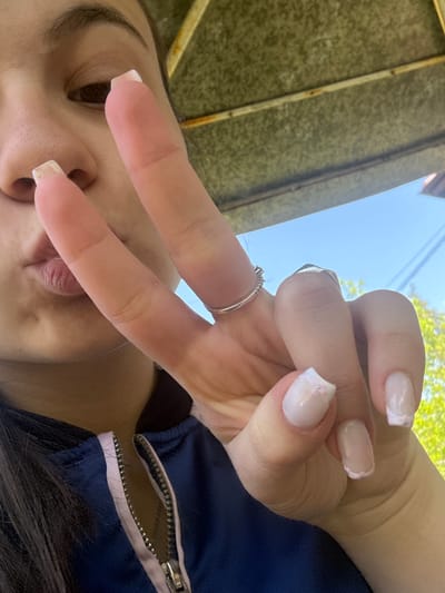 The image is a selfie taken from a low angle, focusing on a person's face and hand making a peace sign. The person has dark hair and light skin. They are wearing a silver ring on their middle finger. Their nails are manicured with a light pink base and white tips with iridescent glitter accents. The person is wearing a blue jacket with a zipper. The background includes a corrugated metal or textured surface, possibly a roof or awning, and a patch of blue sky with some green foliage visible, suggesting an outdoor setting. The location is not definitively discernible as Radomir, Bulgaria from the visual information provided.