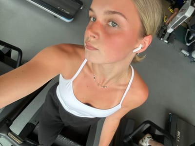 The image captures an inverted, close-up selfie of a young woman taken inside an indoor gym, likely in Whistler, Canada. The woman is shown from the chest up, looking directly into the camera with a neutral expression. She has blonde hair, fair skin with some visible freckles and a flushed appearance on her cheeks and nose, suggesting recent physical exertion. She wears a white spaghetti-strap tank top, black pants, and white wireless earbuds in her ears. Notable accessories include a small silver hoop nose piercing and a delicate silver necklace with small green and white beads. A small, simple tattoo is visible on her upper left arm.
The setting is a gym, identifiable by the dark gray floor and large pieces of exercise equipment. The woman appears to be positioned on or near a leg press machine, with white sneakers featuring black stripes (possibly Adidas) visible on the footrests. Another piece of gym equipment is partially visible in the blurred background to the left. The lighting is consistent with an indoor gym environment. The overall scene depicts a moment during a workout session.