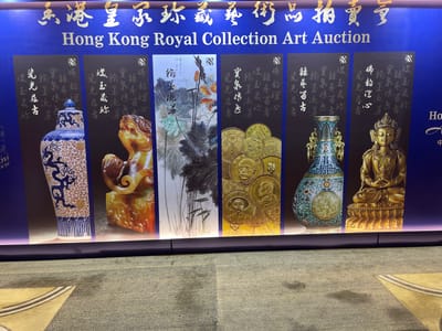 This media file captures a promotional banner for the "Hong Kong Royal Collection Art Auction." The banner is predominantly blue and features an array of artworks and artifacts.
The subjects displayed include a detailed blue and white porcelain vase adorned with dragons, a golden sculpture of a mythical creature, a traditional Chinese ink painting of lotus flowers, a collection of gold coins featuring portraits of historical figures, an ornate cloisonné vase, and a gilded statue of Buddha.
The banner's title, "Hong Kong Royal Collection Art Auction," is prominently displayed in English and Chinese characters. There are also smaller blocks of Chinese calligraphy accompanying each artwork, likely descriptions or titles related to the auction items.
The setting appears to be an indoor exhibition or event space, with a textured grey carpet in the foreground. The lighting is bright, highlighting the rich colors and details of the displayed items. The overall impression is one of elegance and cultural heritage, promoting a high-end art auction. The location context provided is Futian District, China, suggesting this is a promotional display in that area.