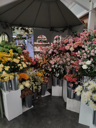 The image depicts an indoor display of artificial flowers, arranged under a gray, gazebo-like structure. The flower selection is abundant and diverse, featuring a range of colors including yellow, orange, white, and various shades of pink and red. The flowers are arranged in metallic buckets which are placed on white rectangular stands. The flooring is a dark gray, and the backdrop reveals glimpses into the surrounding interior, including shelves with additional decorative items. The scene is located in Colchester, United Kingdom.