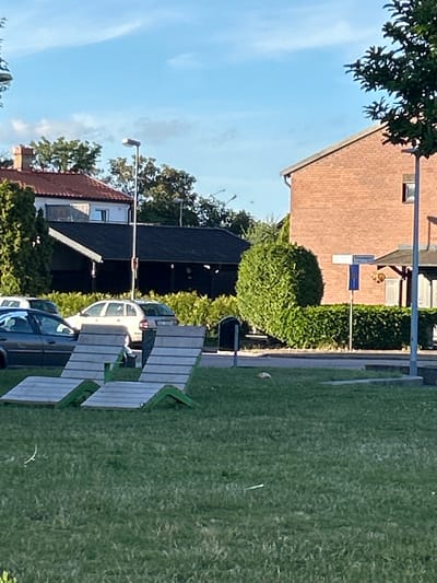 The image depicts a residential scene on a bright, clear day, likely in the late afternoon given the angle of the sunlight casting shadows. The setting appears to be a suburban area in Helsingborg, Sweden, characterized by brick houses with red-tiled roofs and well-maintained green lawns.
In the foreground, a park bench made of light-colored wood slats and green metal framing is prominent, positioned on a paved walkway. Behind the bench, a light-colored car is parked. Further back, a darker car is visible, and a portion of a dark-roofed building is seen, possibly a garage or shed.
The background features a multi-story brick house with a brown roof and visible windows. Adjacent to this house is a tall, dense green hedge, and beyond that, a neatly mowed lawn stretches into the distance. Another house with a red-tiled roof and white siding is visible on the left side of the image, with trees providing a leafy canopy. Streetlights are also present, one clearly visible on the left side of the image. The sky is a clear, bright blue with a few wispy clouds. There are no people visible in the image. The overall impression is one of a peaceful, well-kept residential environment.