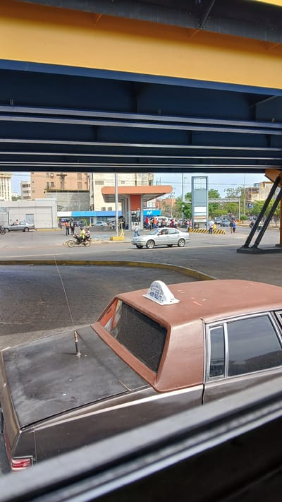 The image, taken in Puerto La Cruz, Venezuela, shows a street scene from under a bridge. A brown and black car is in the foreground, partially obscuring the view. In the background, a gas station with a blue and white "M" logo is visible, with several people nearby. A white car and motorcycles are on the road. Buildings line the street behind the gas station. The bridge's steel structure is in the foreground, painted yellow and blue.