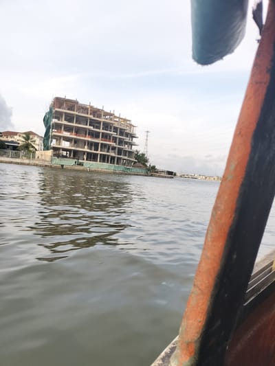 An exterior, eye-level, medium shot captures a partially constructed building on the waterfront in Ikoyi, Nigeria. The building, a concrete structure with visible rebar, is several stories high and appears to be covered in green netting on one side. In the foreground, the dark, rusty edge of a boat or pier obstructs the lower right quadrant of the image, with a portion of the water filling the left side. The water's surface is rippled, reflecting the overcast sky and the building. In the background, other buildings and palm trees are visible along the shoreline, along with a tall electrical pylon. The sky is cloudy and pale.