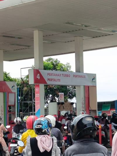 A queue of motorcyclists, wearing helmets of various colors and designs, are lined up at a Pertamina gas station in Cotbadatunong, Indonesia. The station has a canopy supported by concrete pillars, with fluorescent lighting visible on the underside. A sign indicates "4 PERTAMAX TURBO PERTAMAX PERTALITE" along with the Pertamina logo. Another sign on a pillar shows a red arrow with "JALUR KHUSUS" (Special Lane). Trees are visible behind the station. The motorcyclists are mostly facing away from the camera, waiting to refuel. The scene appears to be during daylight hours.
